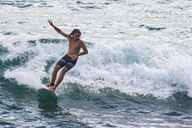 A shirtless surfer in dark boardshorts carving a graceful turn across a small, foamy wave at Noosa Beach.