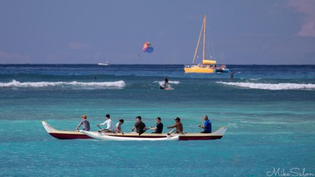 A long white outrigger canoe filled with paddlers slicing through bright blue ocean water with a yellow catamaran in the background under a clear sky.