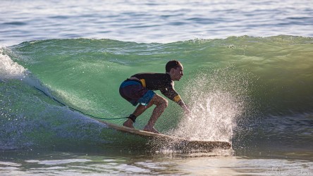 A surfer crouched low on a surfboard riding along a glassy green wave illuminated by early morning light at Noosa.