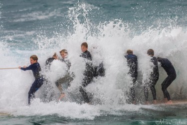 A group of boys in wetsuits holding onto a rope boundary marker at Bronte Ocean Pool while a huge wave crashes directly over them in a wall of white spray.