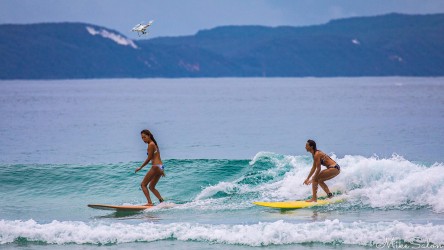 Two women in bikinis riding a small wave side-by-side on longboards while a white drone flies overhead against distant headland hills.