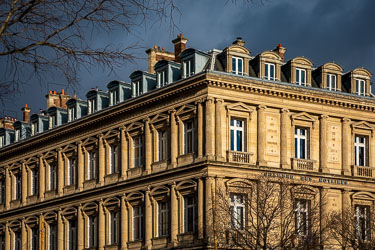 An angled low-angle shot of the classical stone façade of the Chambre des Notaires at Place du Châtelet in Paris under a dark, stormy grey sky. The warm sunlight illuminates the limestone building, highlighting fluted columns, detailed window frames, ma
