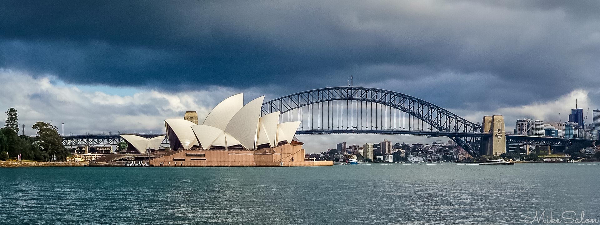 A dramatic perspective of Sydney Harbour, showcasing two of Australia's most recognized architectural landmarks framed against a brooding sky. In the foreground, the gleaming white sails of Jørn Utzon A dramatic perspective of Sydney Harbour, showcasing two of Australia's most recognized architectural landmarks framed against a brooding sky. In the foreground, the gleaming white sails of Jørn Utzon