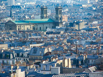 View over Paris rooftops with chimneys and slate roofs under pale blue light, showing the city