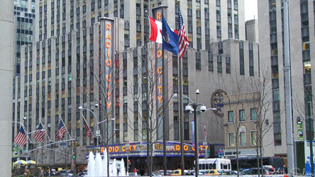 Exterior of Radio City Music Hall in Rockefeller Center, New York City, with tall Art Deco façade, flags, and street scene.