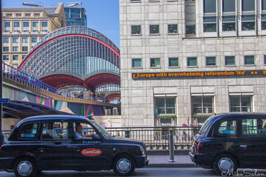 Canary Wharf DLR station in London with arched glass roof, modern office buildings, and black taxis in foreground.