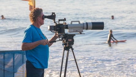 Cameraman using a large telephoto video rig on a tripod at Noosa Beach, filming surfers in the late‑afternoon light.