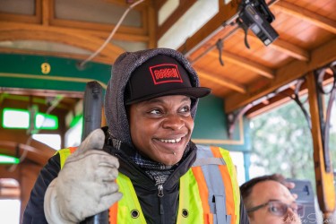 Smiling cable‑car operator in a yellow safety vest and cap, driving a wooden San Francisco cable car on a chilly day.