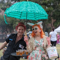Two cheerful festival goers at the Surry Hills Festival in Sydney, one holding iced tea and the other a turquoise umbrella and donation bucket.