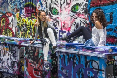 Two young women sitting on graffiti‑covered bins in Hosier Lane, Melbourne, chatting against a vivid mural backdrop.