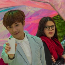 Young man taking a selfie with a smartphone at Kensington Gardens, London, while a woman under a pink umbrella unexpectedly appears in the frame.