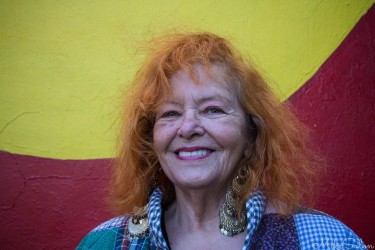 Smiling woman with red hair standing in front of a large Aboriginal flag mural in Newtown, Sydney.