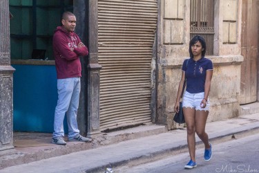 Street scene in Havana showing a man standing near a shuttered shop doorway and a woman walking past, outside a ration store that distributes essentials through Cuba
